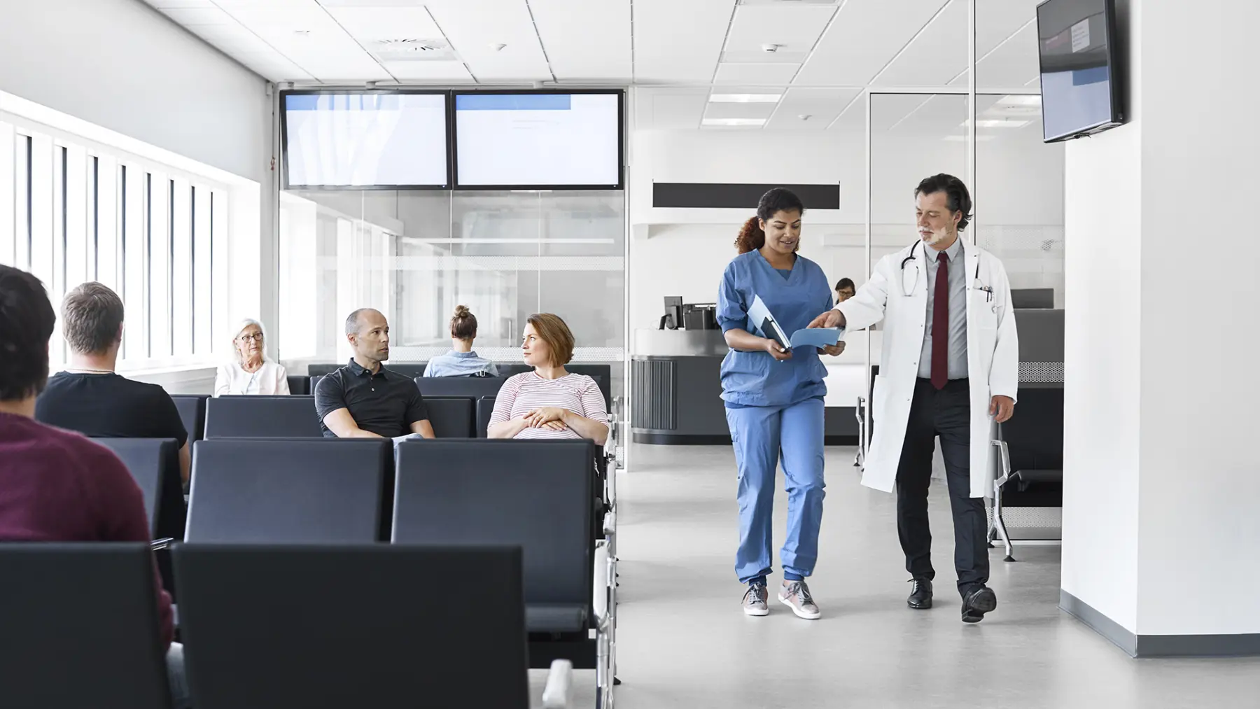 A nurse in scrubs walks with a doctor in a white coat, discussing a patient file in a modern hospital waiting area.