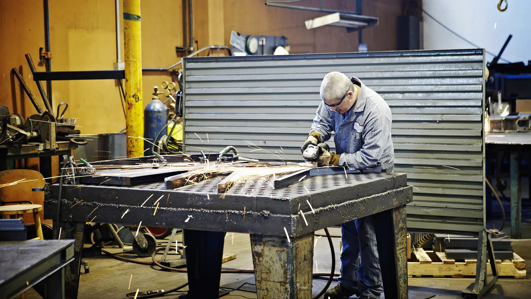 A worker in protective gear uses a grinder on a metal workpiece, surrounded by tools and sparking metal shavings in a workshop.