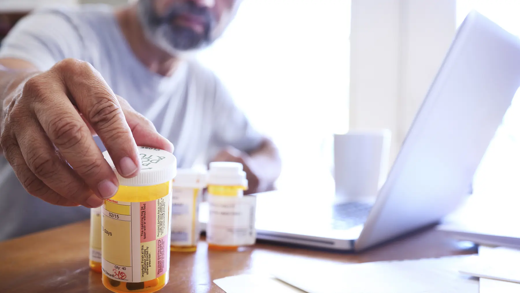 A person reaches for a prescription bottle on a wooden table, with other bottles and a laptop visible in the background.