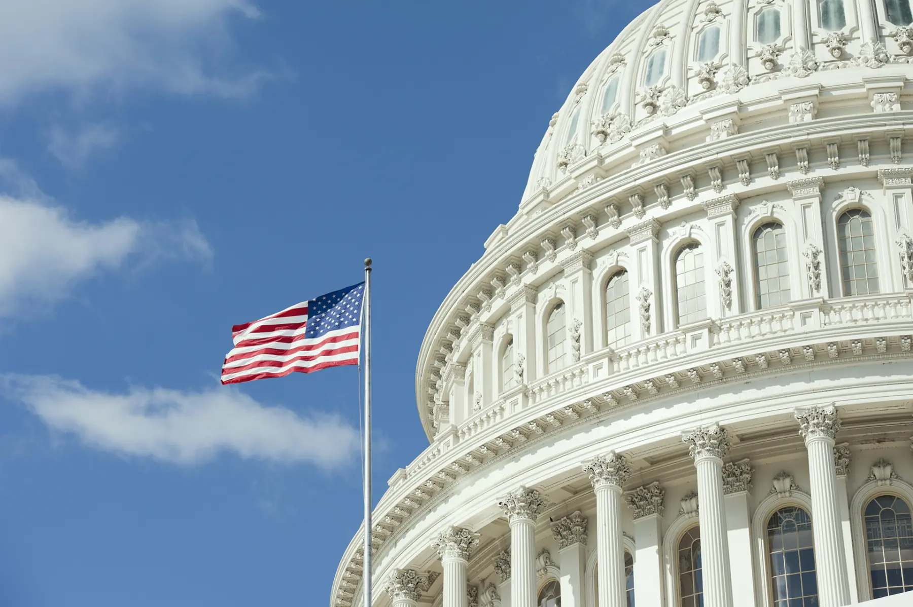 The U.S. Capitol dome partially captures the bright blue sky, with the American flag waving in the foreground.