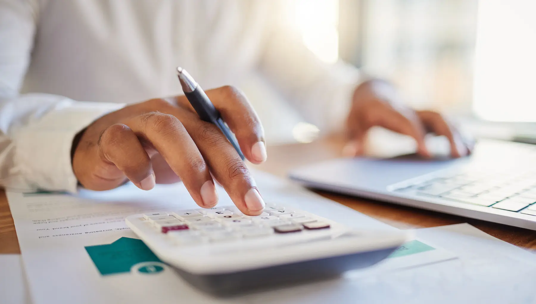 A hand uses a calculator on a wooden desk, while another hand types on a laptop; documents are spread out beneath.