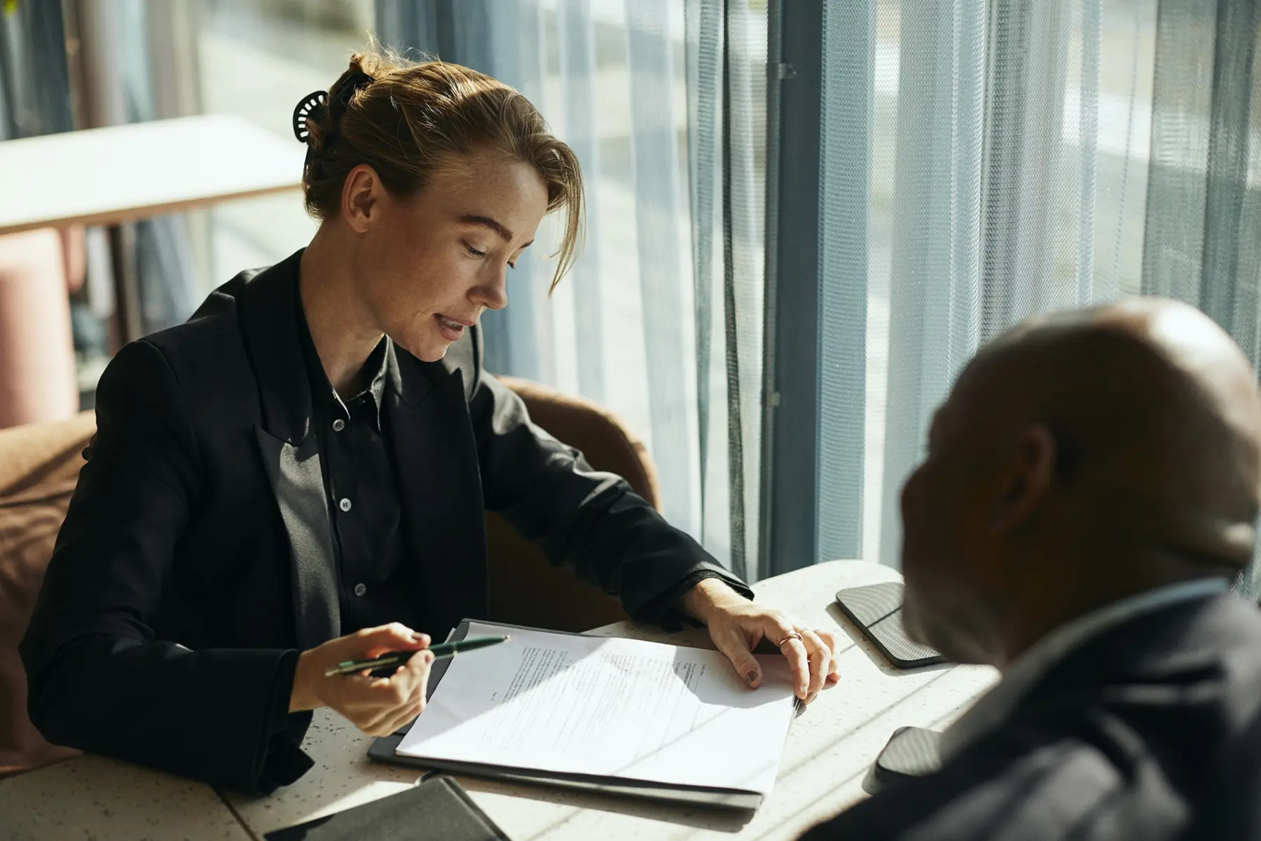 A woman in a black blazer sits at a table, reviewing documents with a serious expression, while a man listens attentively.