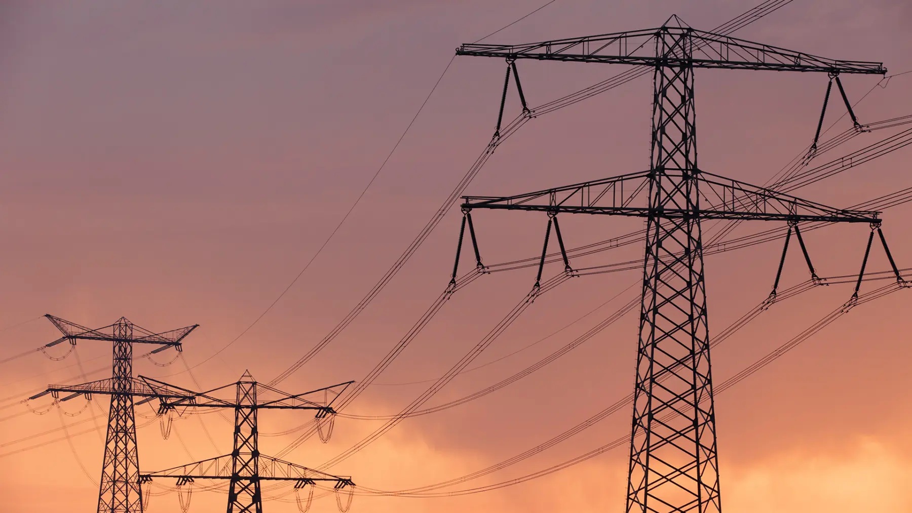 Silhouetted power lines stretch across a colorful sunset, with towers standing tall against a gradient of pink and orange hues.