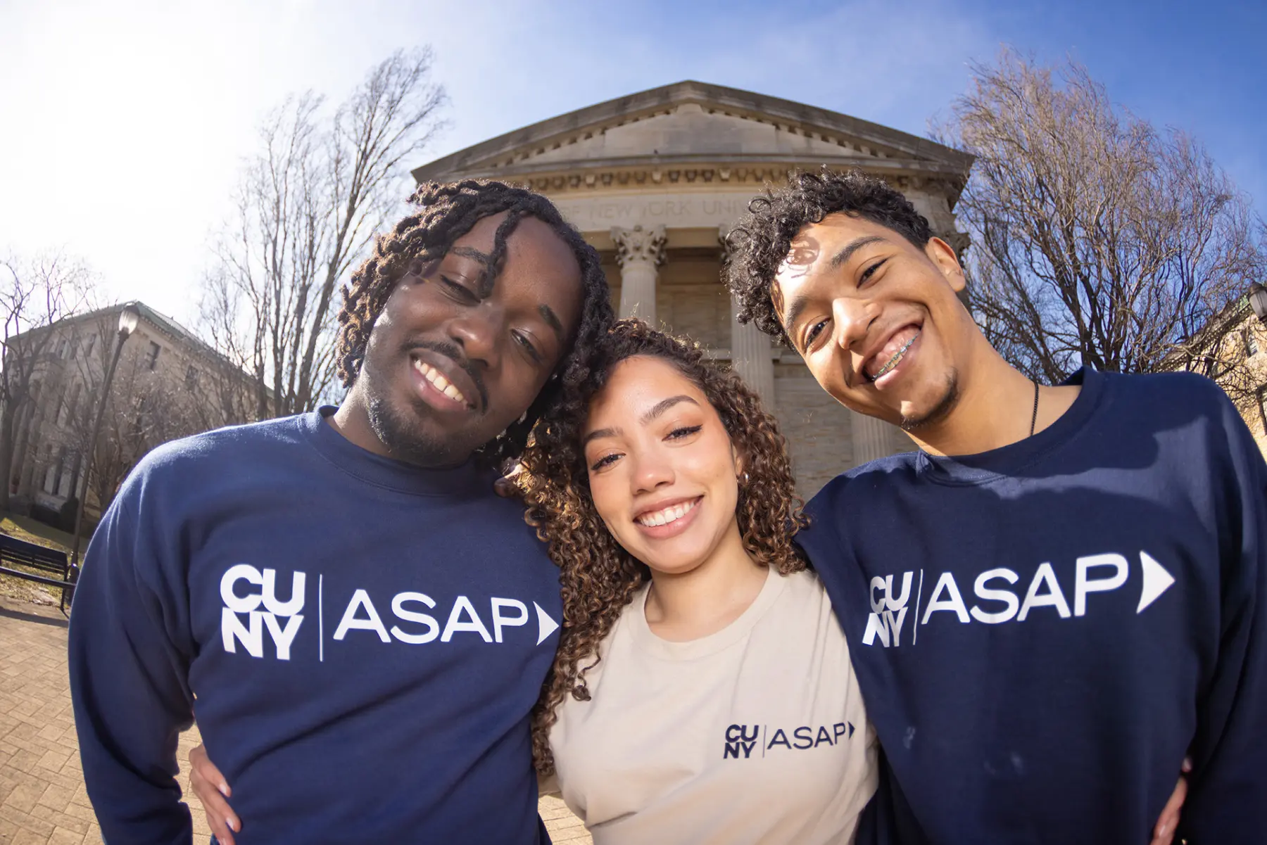 Three smiling young adults pose together outdoors, wearing matching CUNY ASAP sweatshirts, with a historic building in the background.