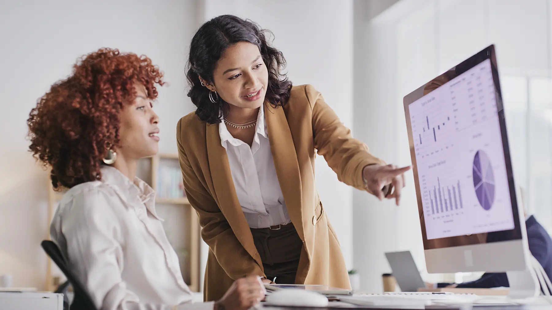 A businesswoman in a brown blazer points at data graphs on a computer screen while discussing insights with a colleague.
