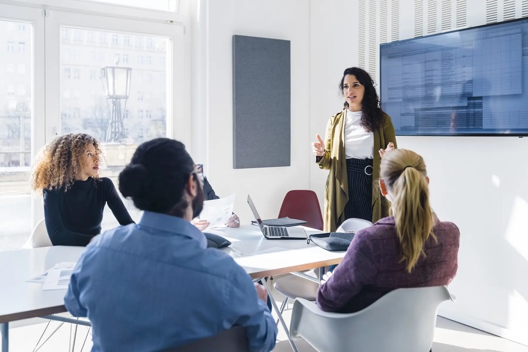 A diverse group in a bright conference room listens attentively as a woman presents information, with a laptop and documents on the table.