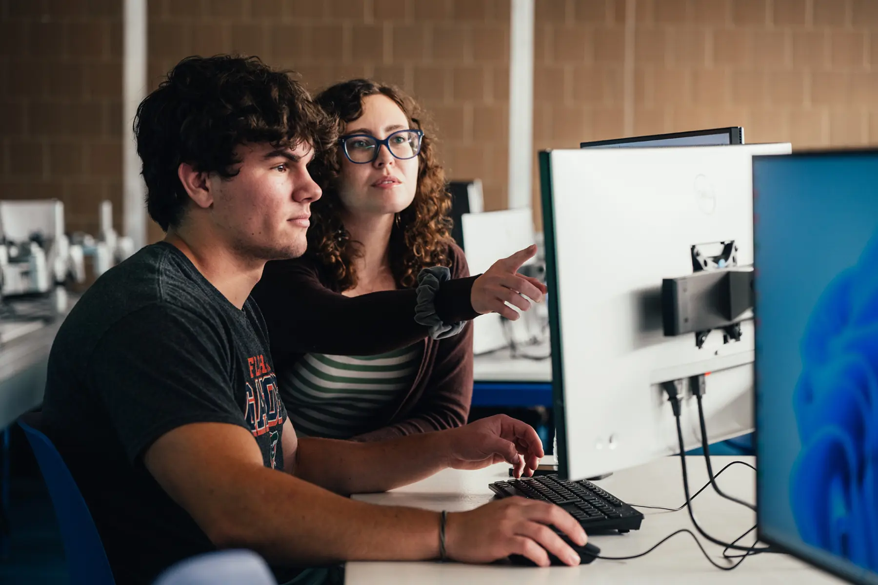 A young woman gestures while explaining something to a young man at a computer, both focused on the screen in a classroom setting.
