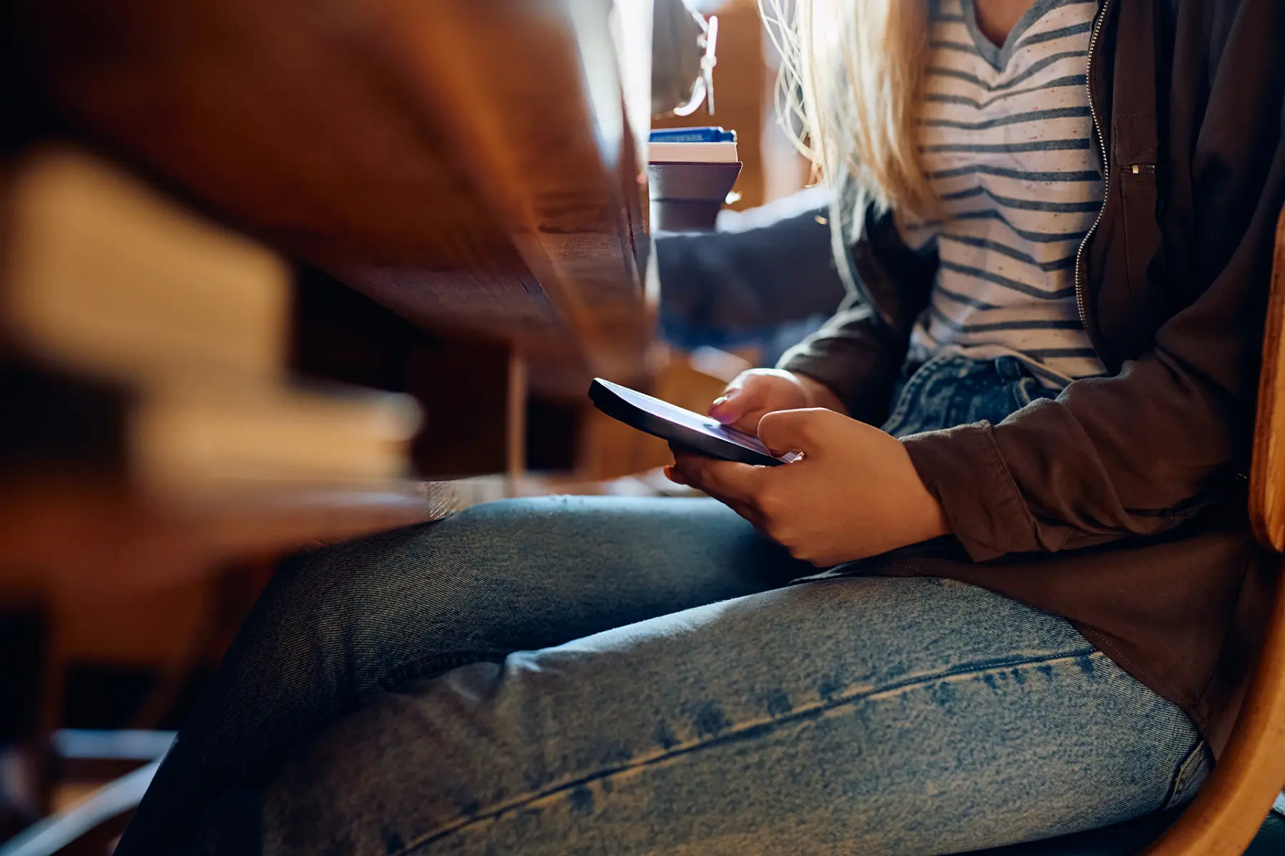 A person sits at a wooden table, holding a smartphone, wearing a striped shirt and denim jeans, with warm lighting enhancing the scene.