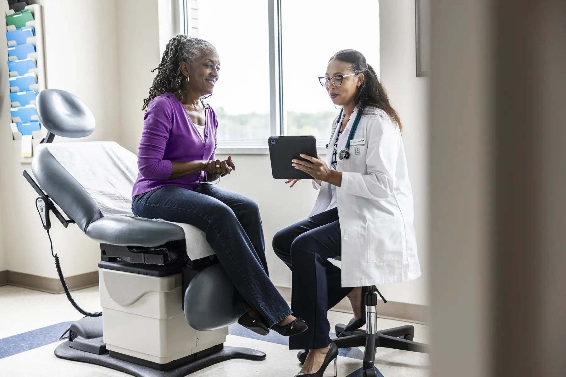 A smiling elderly patient in a purple sweater sits on an exam table, discussing health matters with a doctor using a tablet.