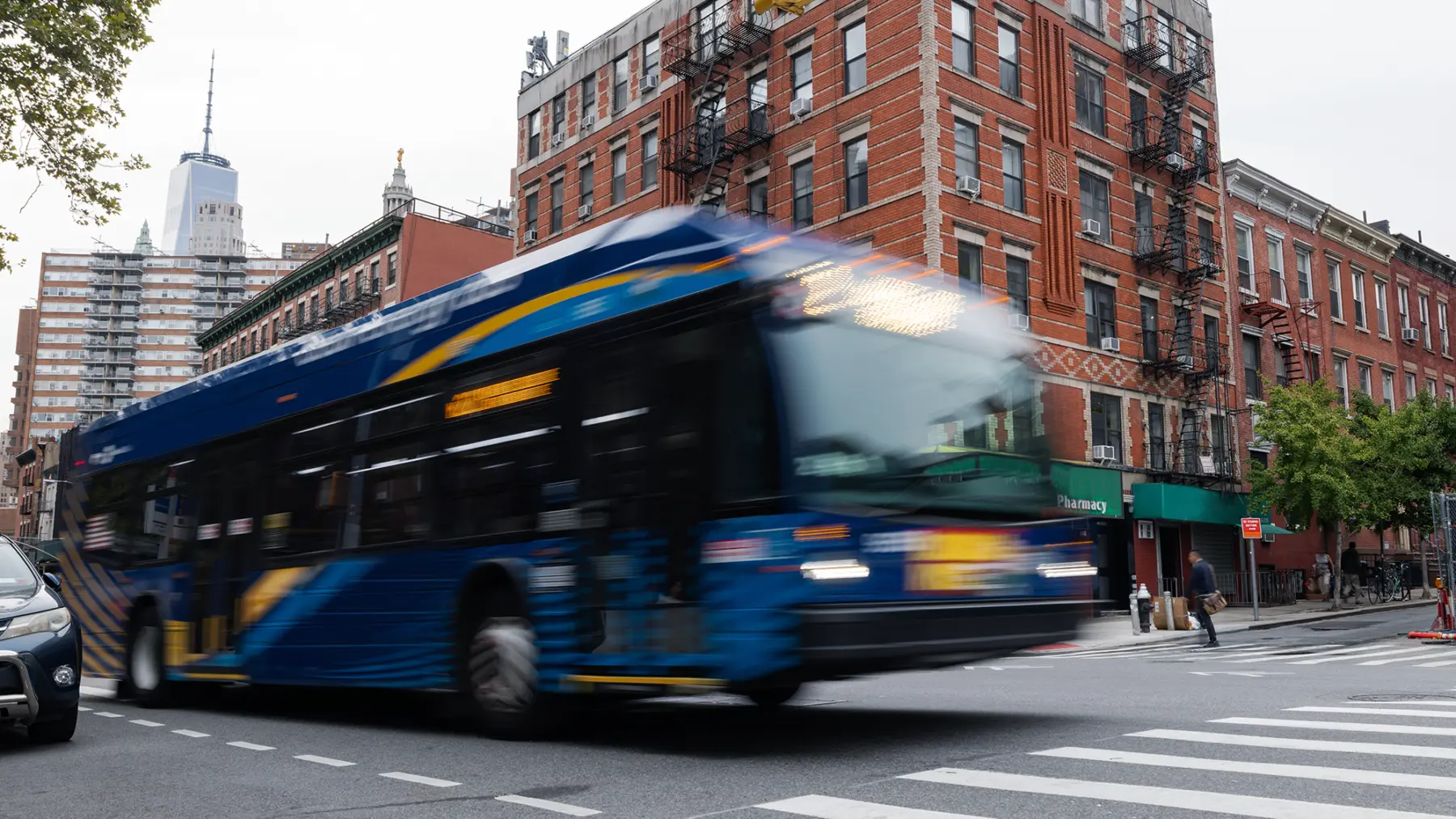 A blue bus speeds through an urban intersection, with red-brick buildings and tall skyscrapers in the background, under a cloudy sky.