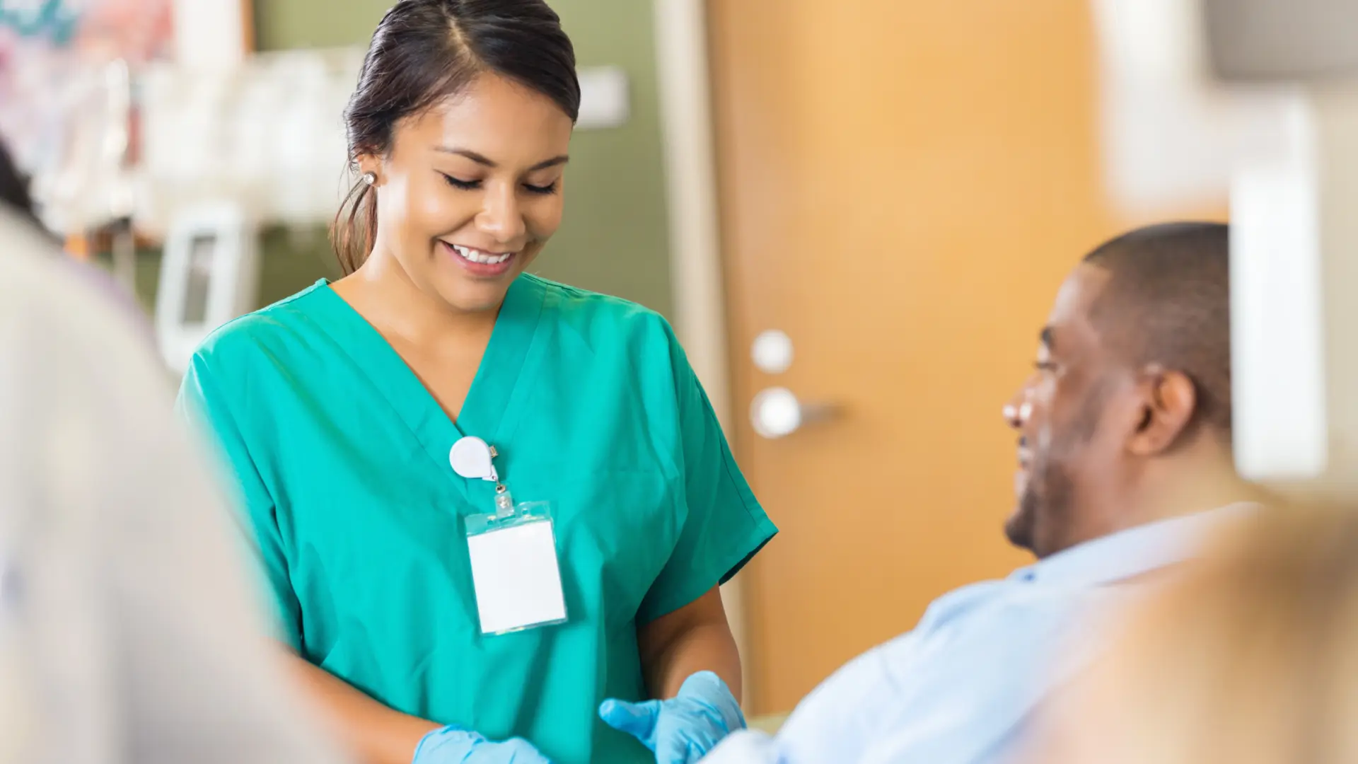 A healthcare professional in green scrubs smiles while preparing to assist a patient sitting comfortably in a medical setting.