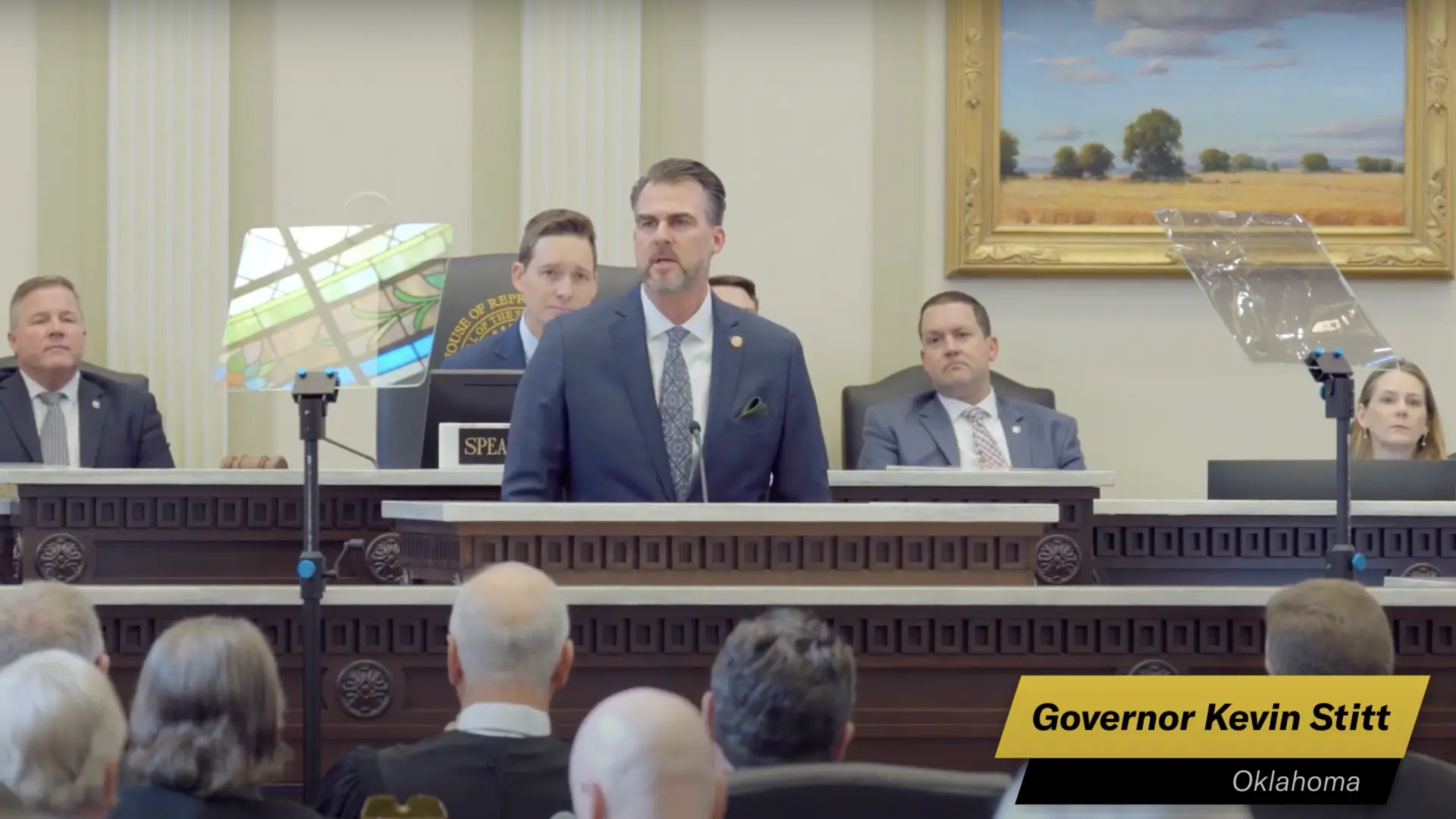 Governor Kevin Stitt stands at a podium speaking in a legislative chamber, with attentive audience members seated behind him.