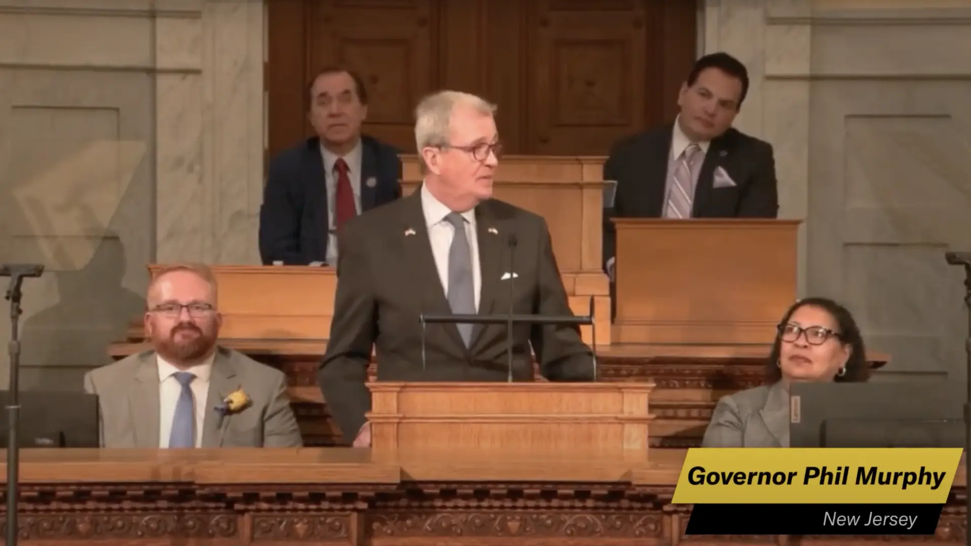 Governor Phil Murphy speaks at a podium in a formal setting, with several attendees, including seated individuals, in the background.