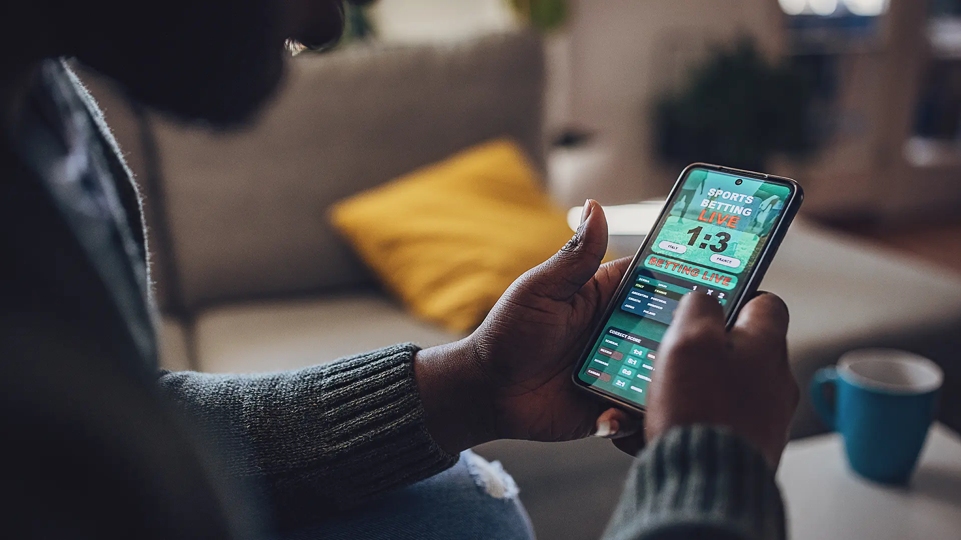 A person is using a smartphone to engage with a sports betting app, while seated on a couch, with a coffee cup nearby.