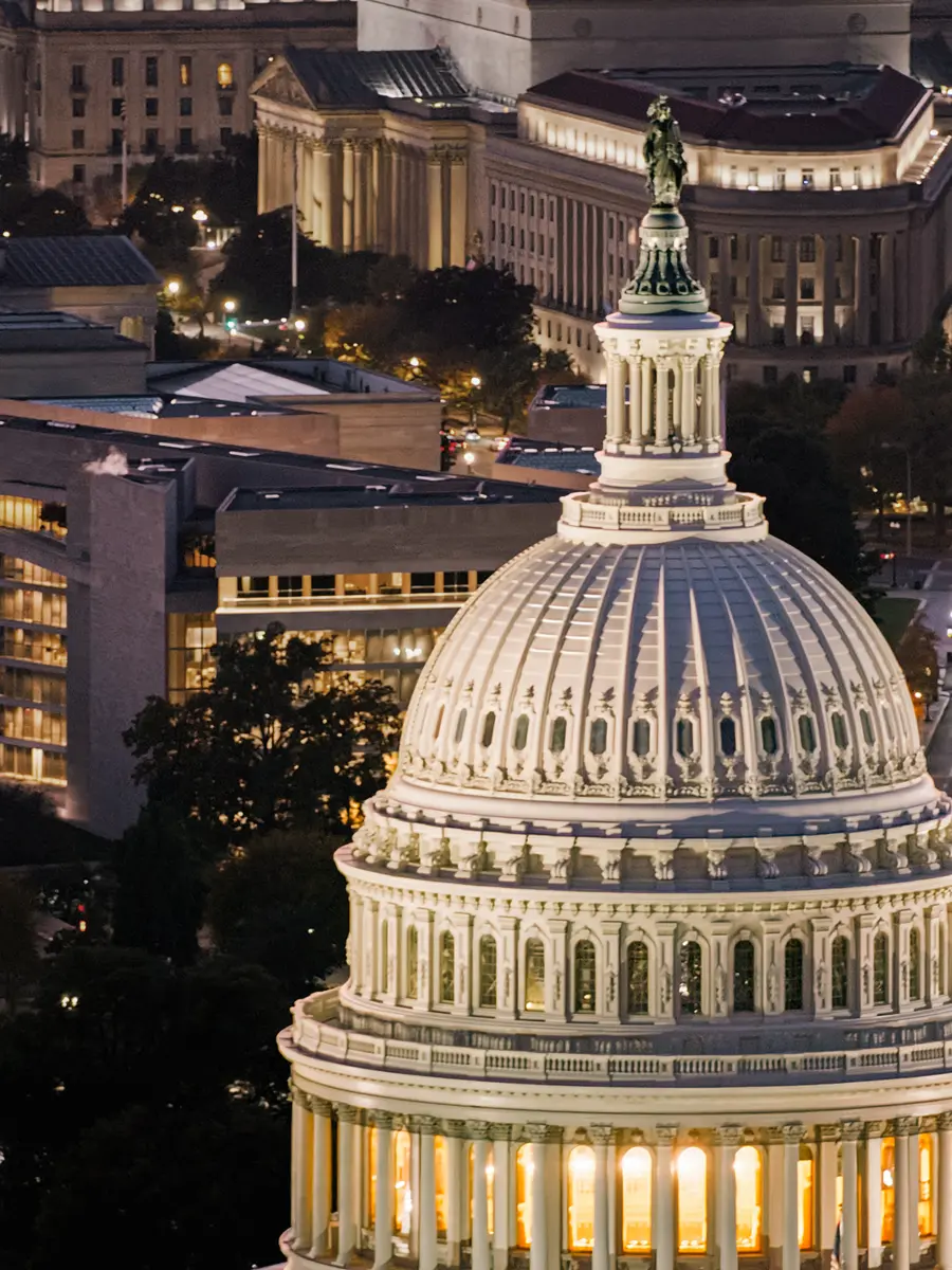 The illuminated dome of the U.S. Capitol stands prominently at night, with surrounding buildings softly lit in the background.