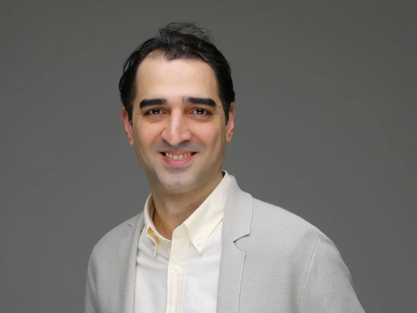 A smiling man with dark hair stands against a gray background, wearing a light gray blazer over a cream-colored shirt.