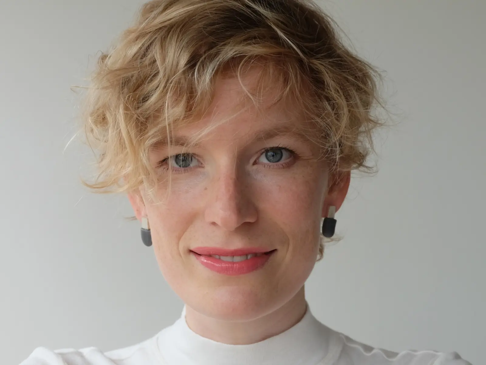 A smiling person with short, curly blonde hair wears a white turtleneck and black geometric earrings, against a light background.