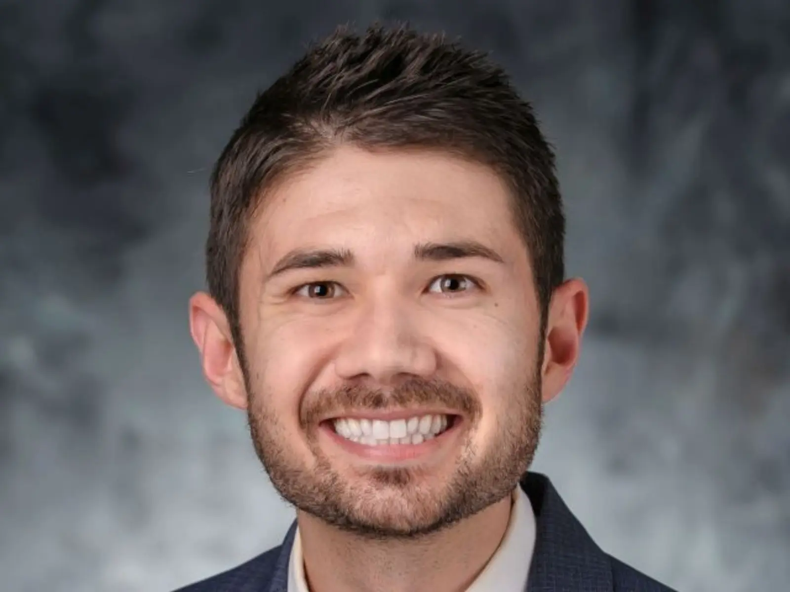 Smiling man with short dark hair, wearing a blue suit and tie, against a soft gray backdrop, exuding a professional and approachable demeanor.