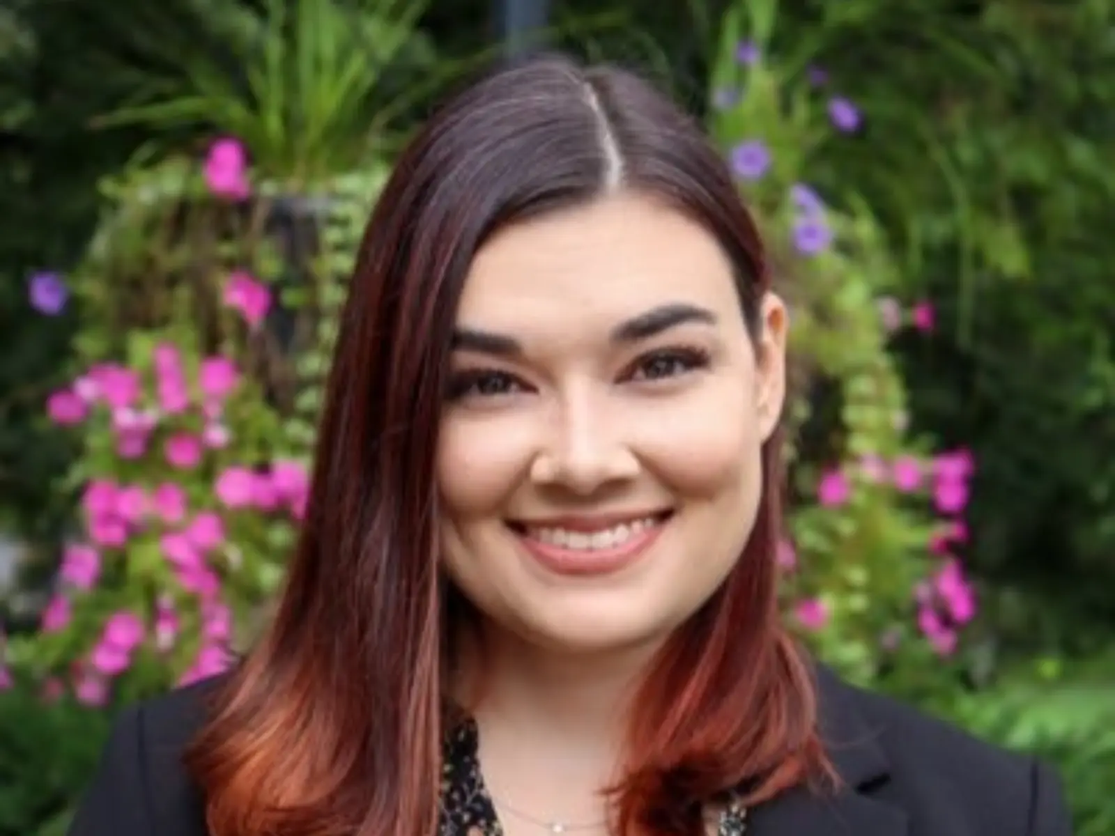 A woman with shoulder-length, brown hair smiles warmly, dressed in a black blazer over a floral top, with vibrant flowers in the background.