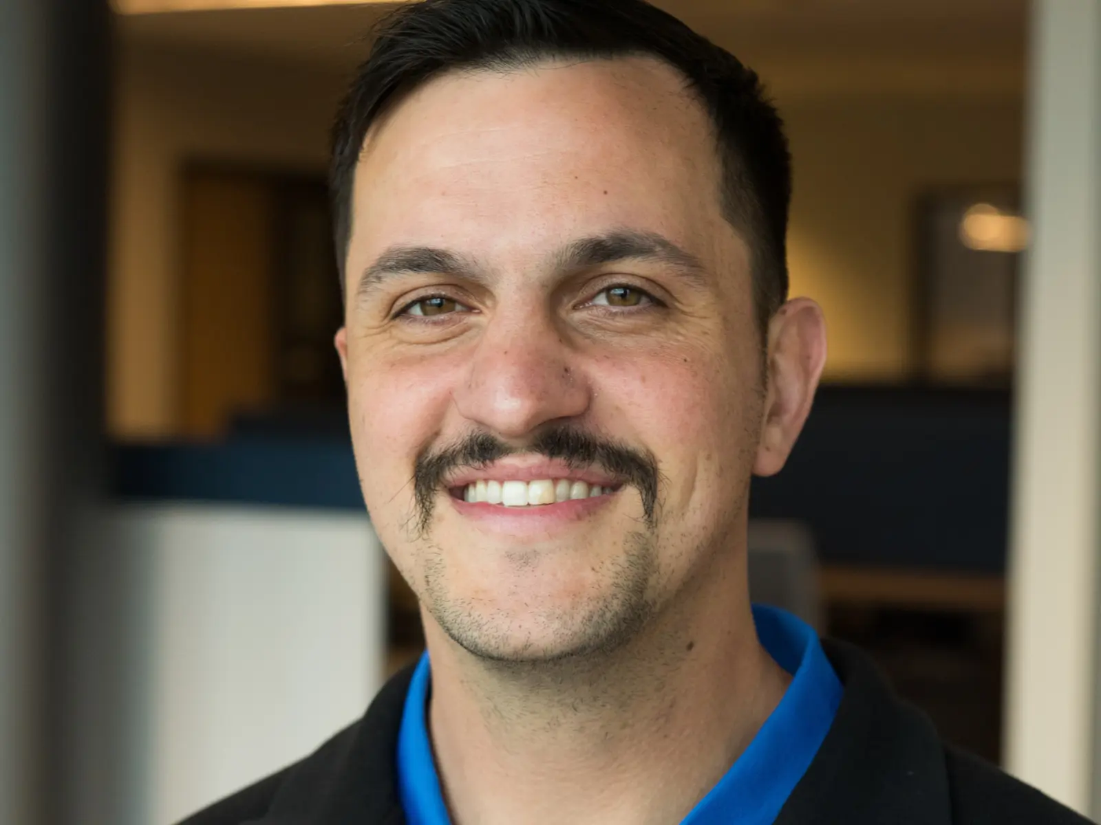 Smiling man with a mustache wears a black jacket over a colorful shirt, standing in an office environment with blurred background.