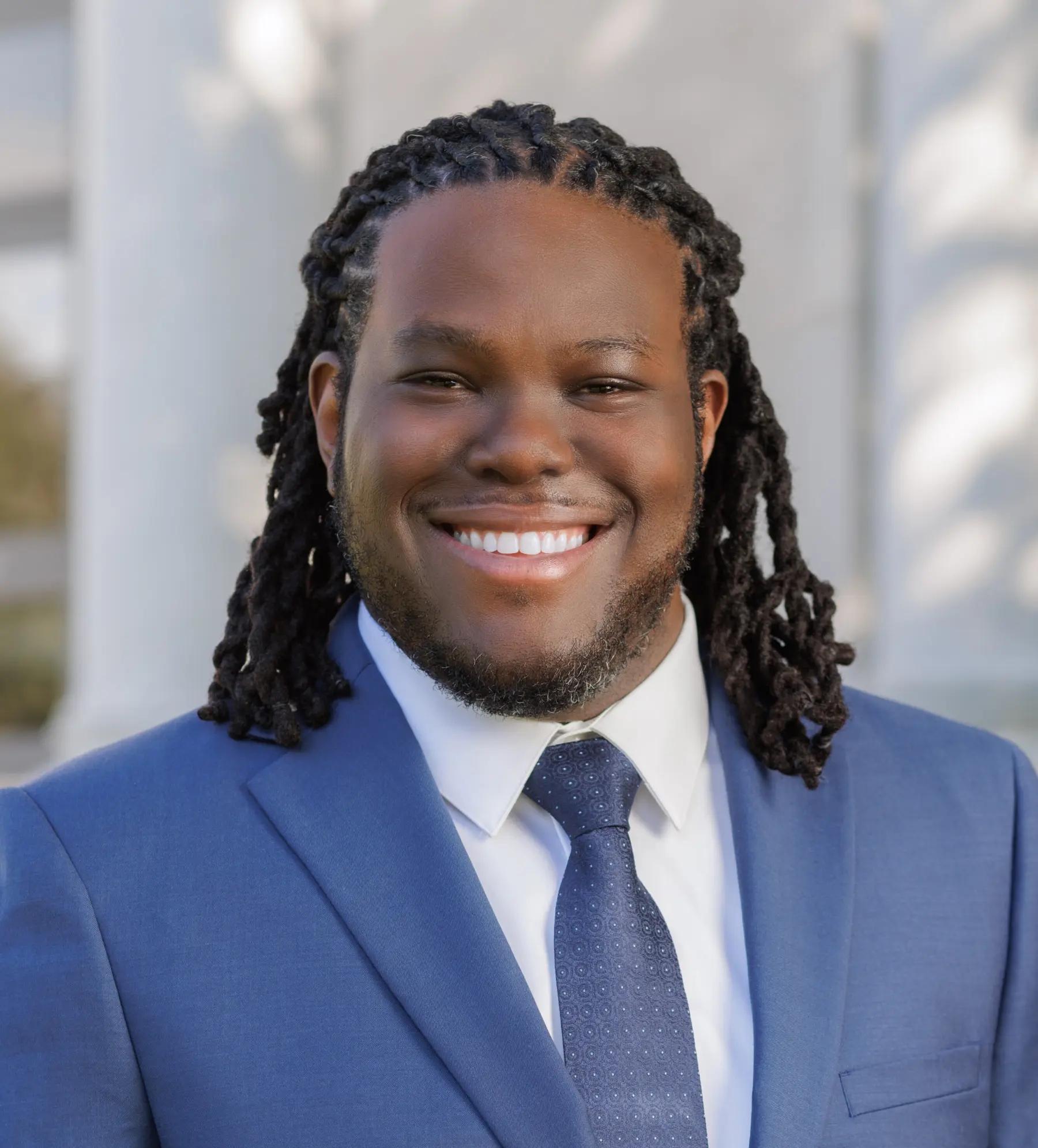 A man with long, dark dreadlocks smiles warmly, wearing a blue suit and patterned tie, against a softly blurred outdoor background.