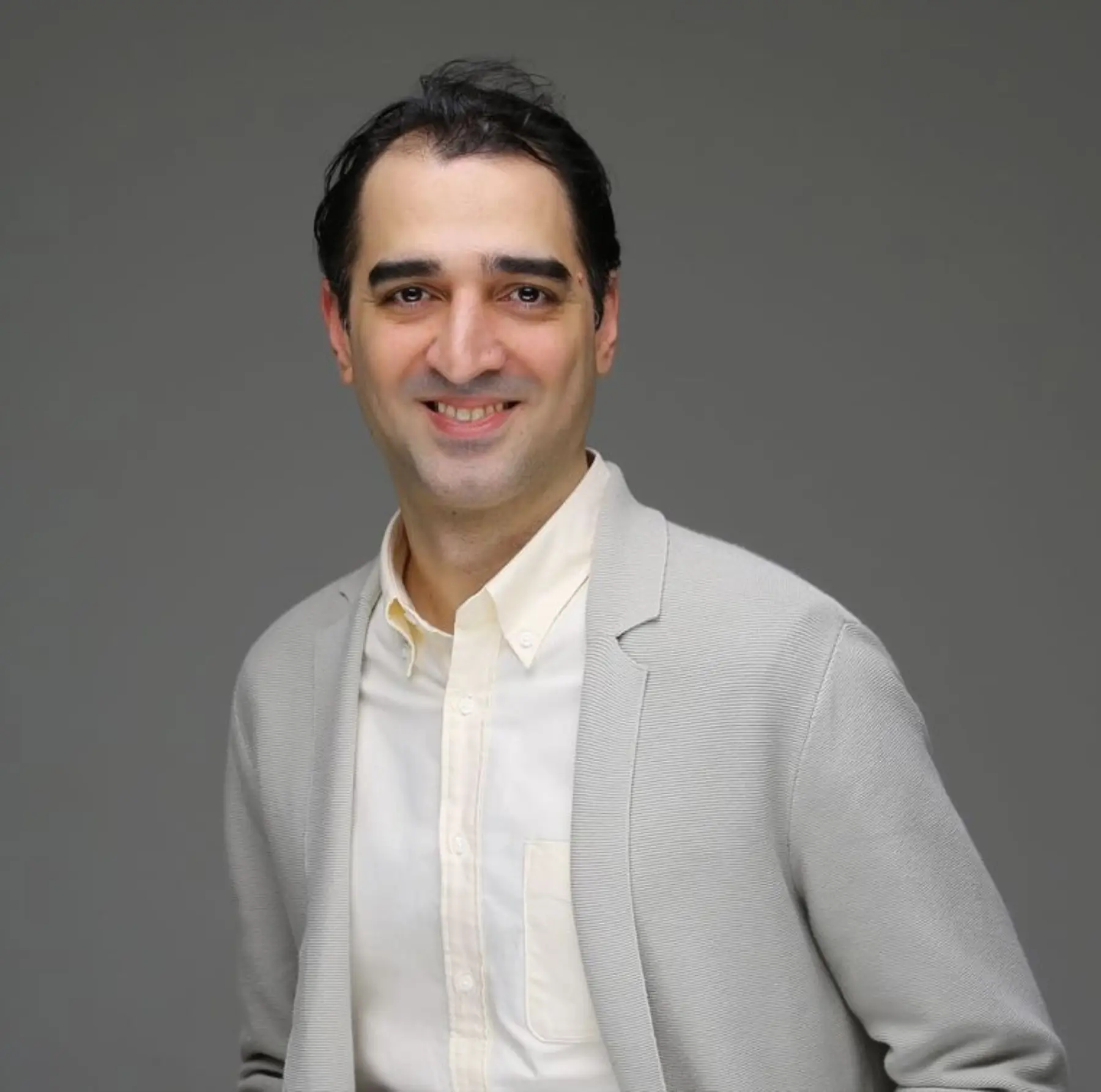 A smiling man with dark hair stands against a gray background, wearing a light gray blazer over a cream-colored shirt.