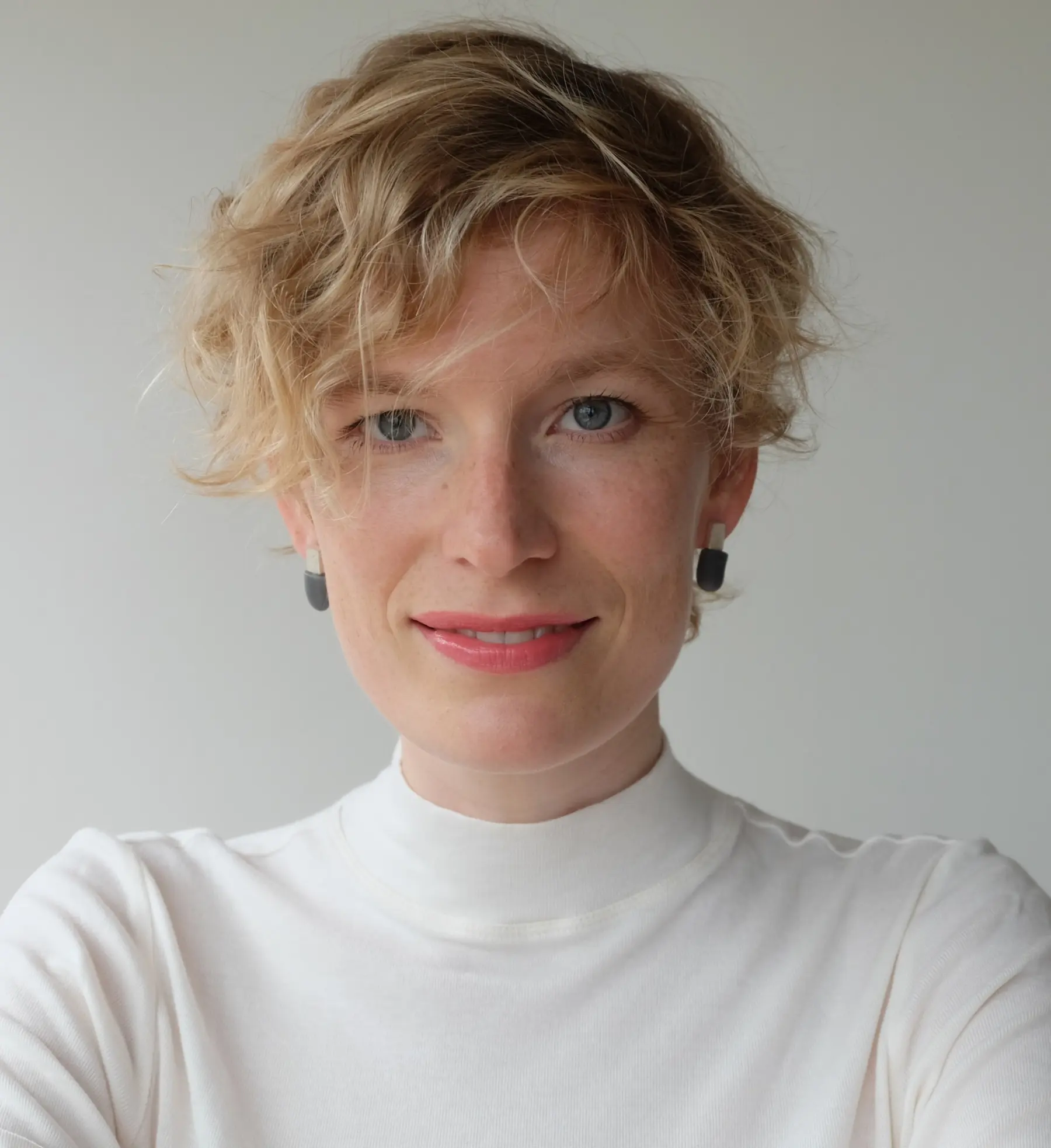 A smiling person with short, curly blonde hair wears a white turtleneck and black earrings, against a light background.