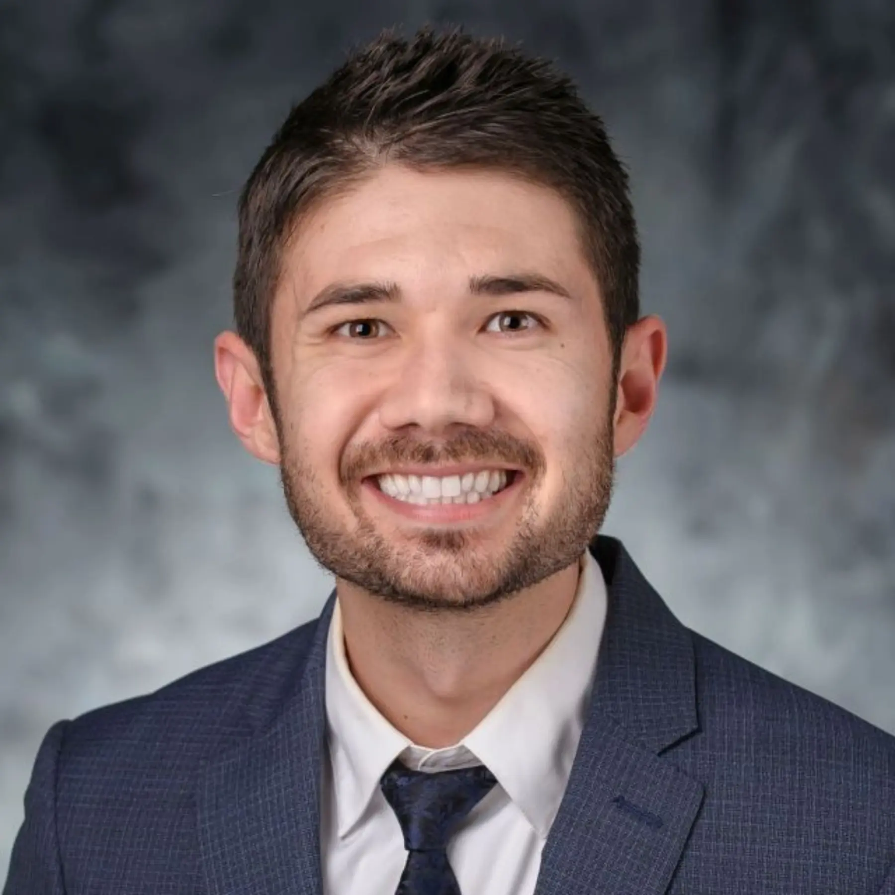 Smiling man with dark hair, wearing a blue suit and tie, against a soft gray backdrop.