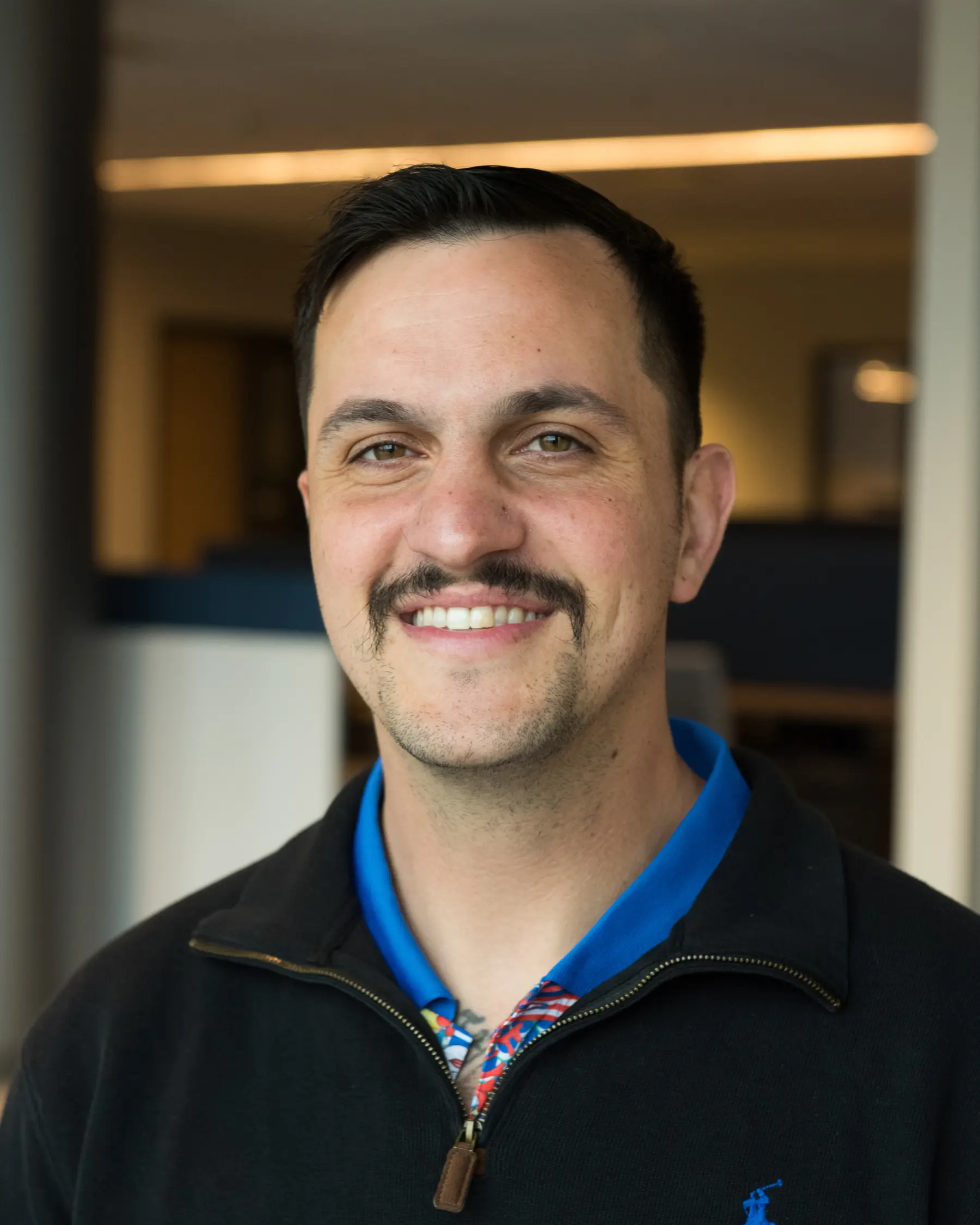 Smiling man with a mustache wears a black jacket over a colorful shirt, standing in an office environment with blurred background.