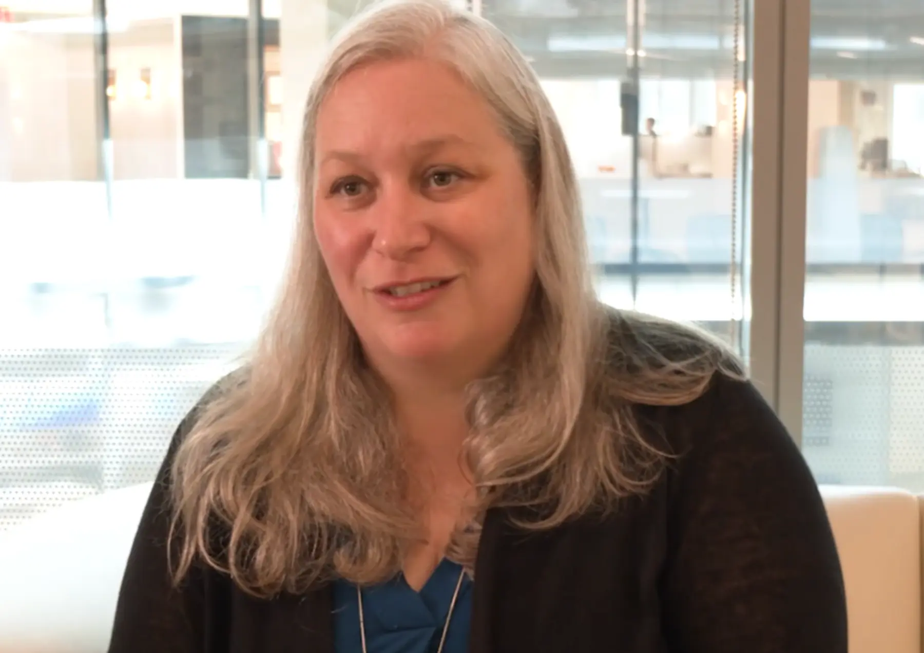 A woman with long, light hair smiles and speaks, seated in a bright, modern office space with large windows in the background.