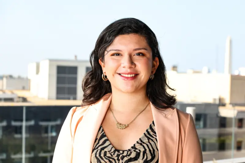 Smiling young woman with curly hair, wearing gold hoop earrings and a delicate necklace, poses against a city backdrop.