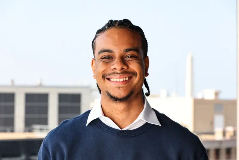 Smiling man with braided hair wearing a navy sweater over a white shirt, standing against a bright sky and urban background.