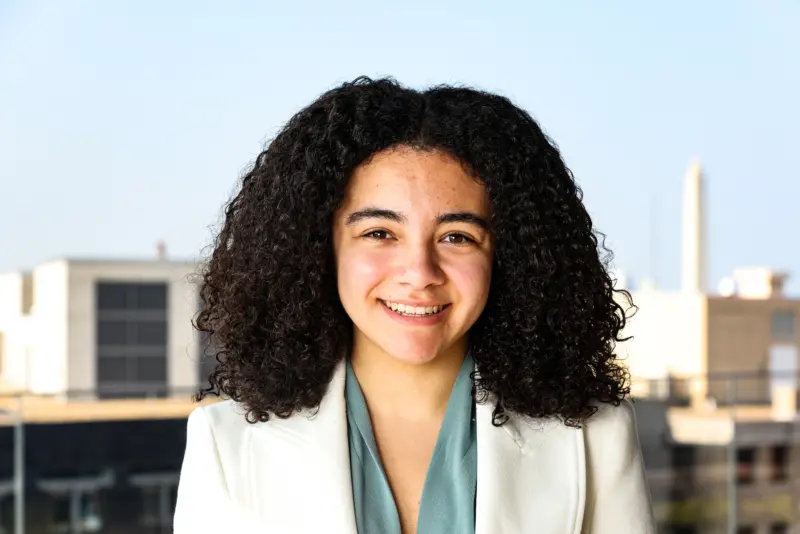 A young woman with curly hair smiles warmly, wearing a white blazer over a light blue top, with a blurred cityscape in the background.
