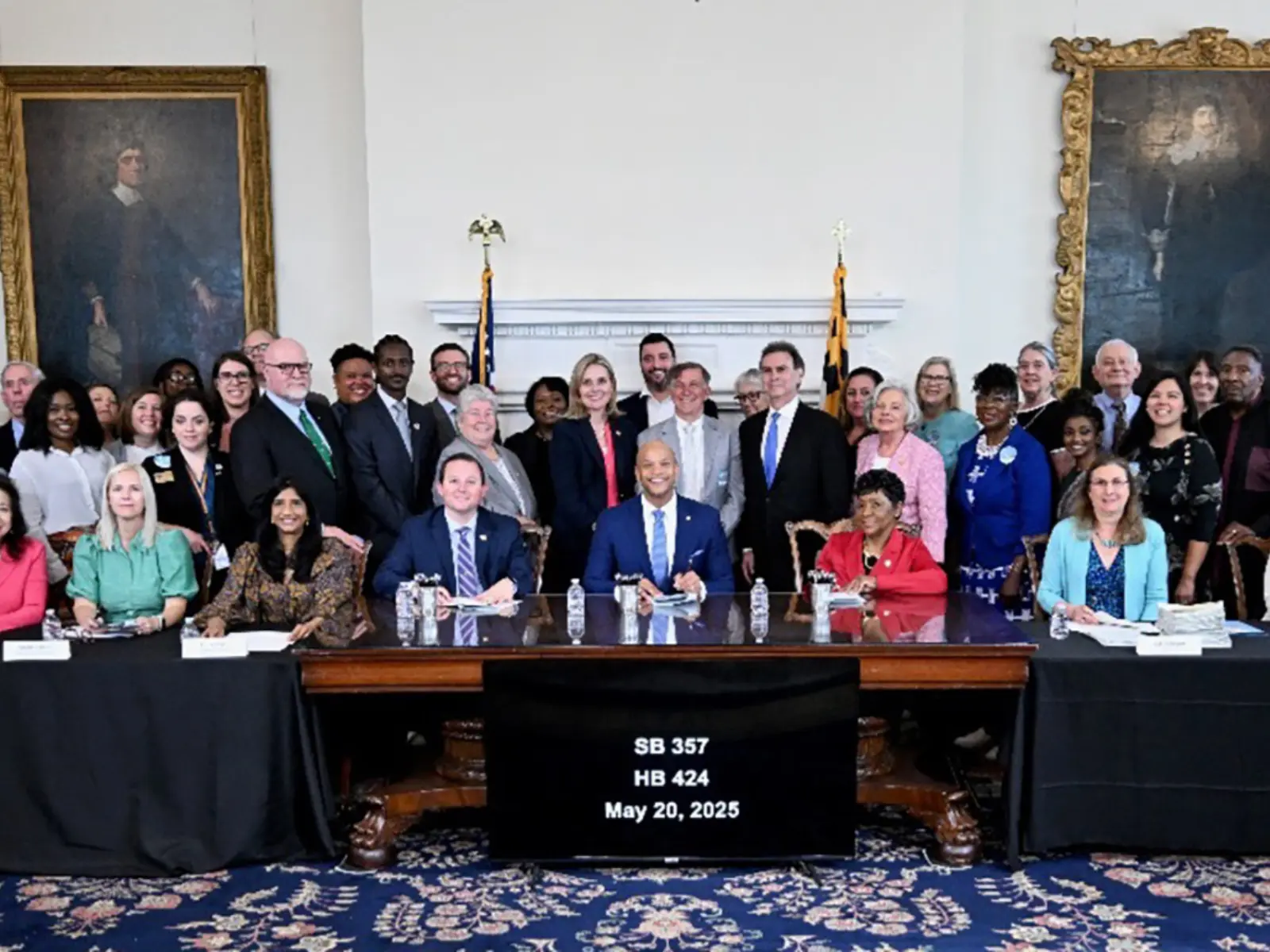A diverse group of officials and supporters gather around a table, celebrating the signing of legislation on May 20, 2025.
