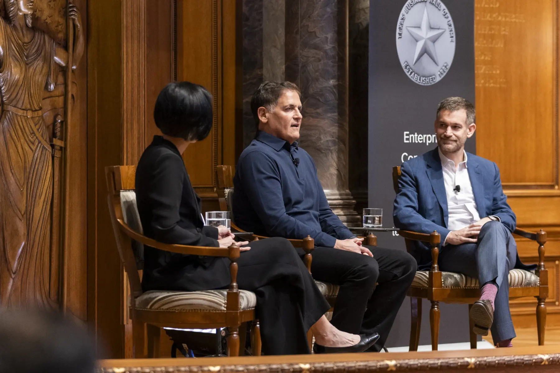Three individuals engage in a discussion on stage, seated in elegant chairs, with a backdrop of detailed wooden architecture and branding.