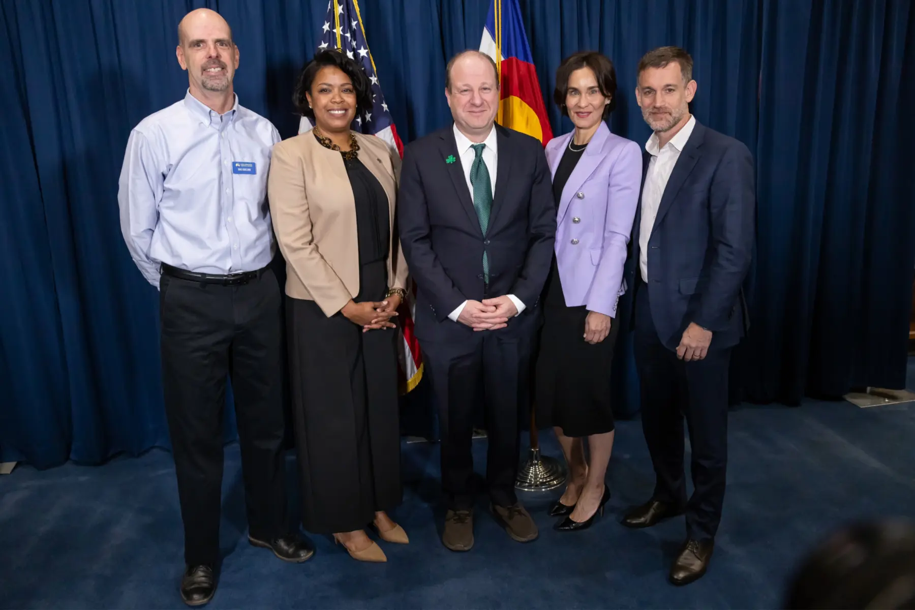 Five professionals pose together in front of a curtain, including an American flag and a Colorado flag, smiling warmly.