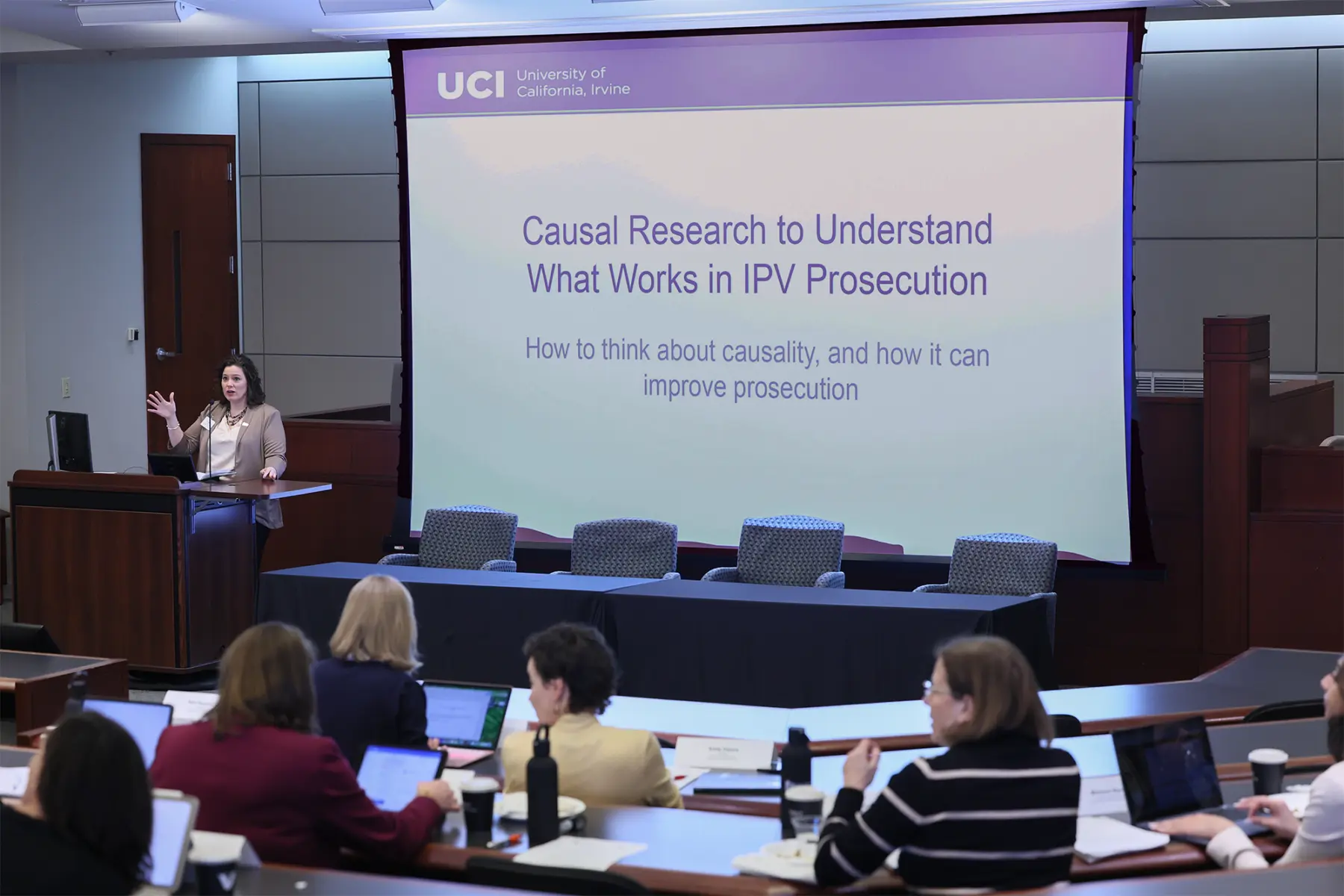 A presenter stands at a podium explaining the role of causal research in improving IPV prosecution, with attendees engaged.