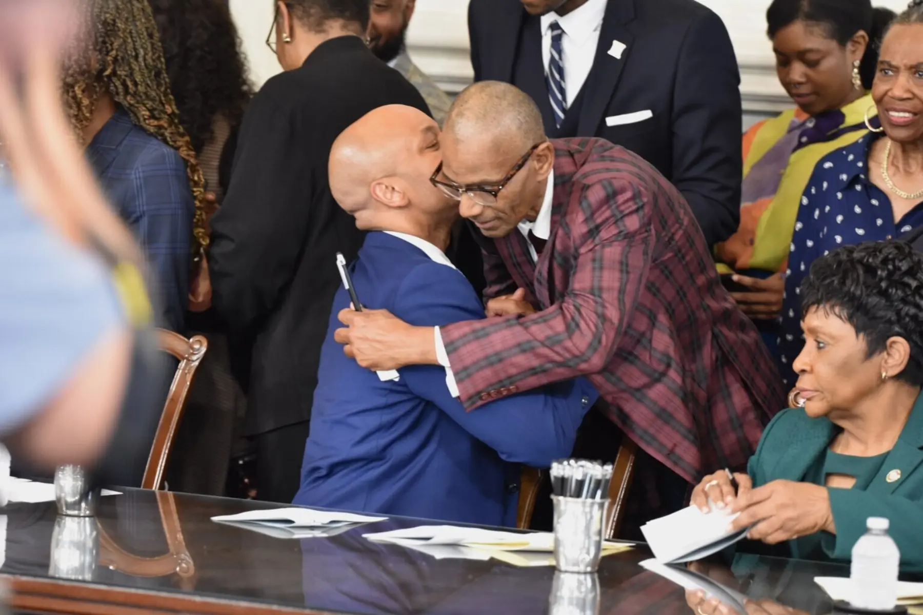 Two men embrace warmly at a formal event, surrounded by attendees. One man is dressed in a blue suit, the other in a plaid jacket.