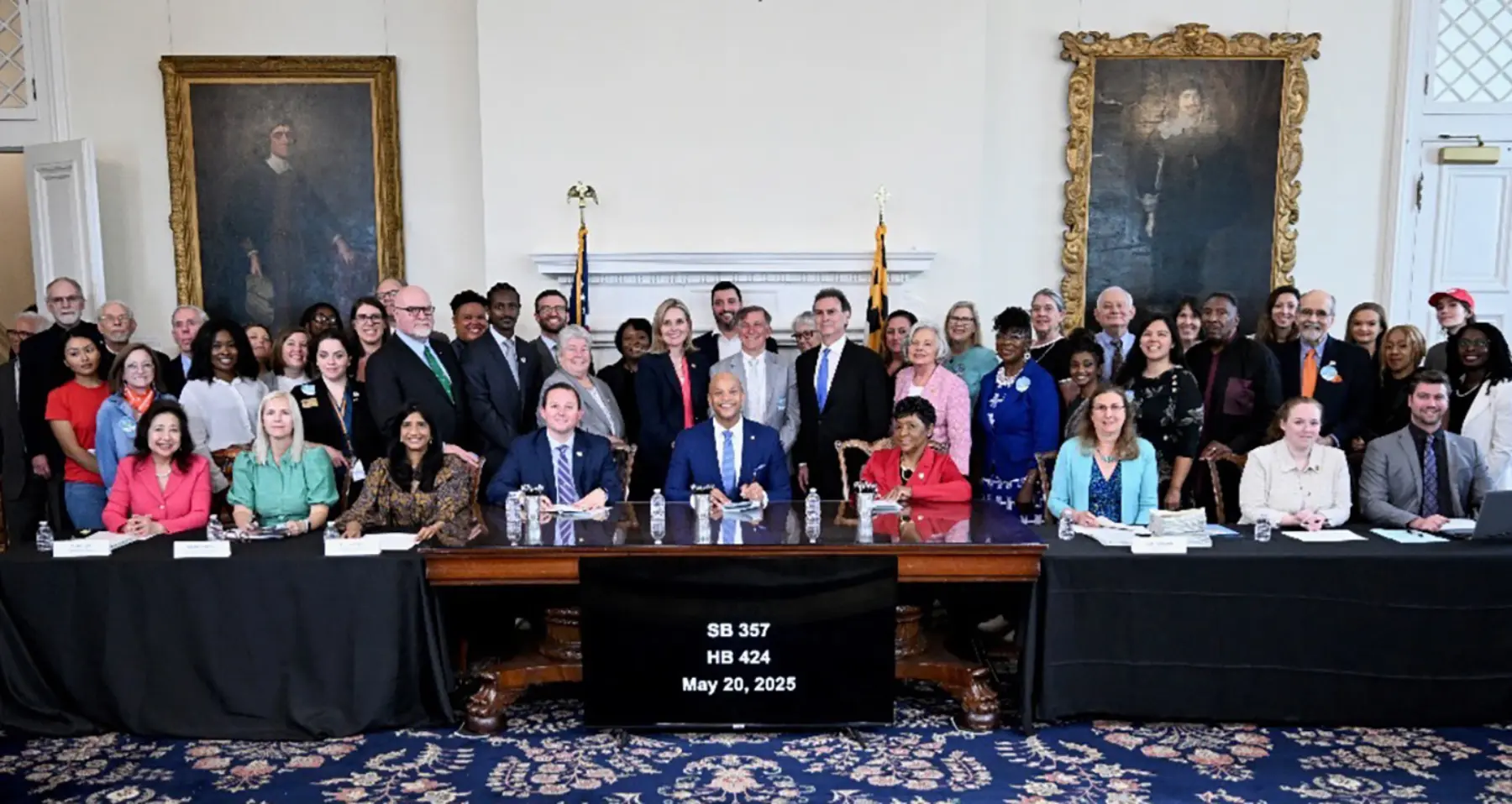 A diverse group of officials and supporters gather around a table, celebrating the signing of legislation on May 20, 2025.