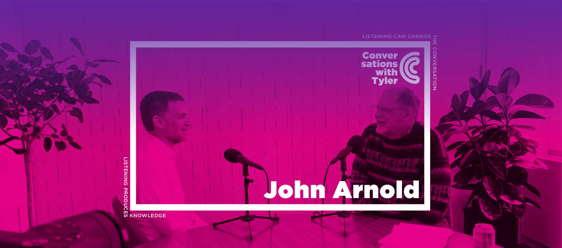 Two men engage in a conversation at a table, microphones in front of them, surrounded by vibrant purple hues and indoor plants.