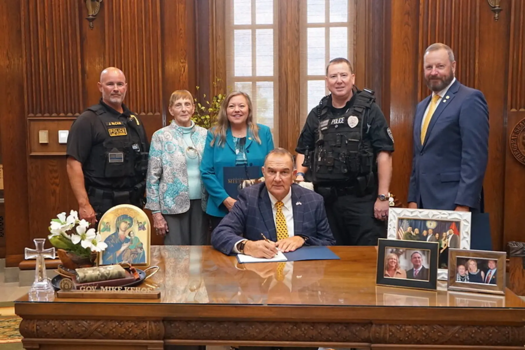 A group of six individuals gathers around a desk in an office, with one person signing documents while others stand nearby, smiling.
