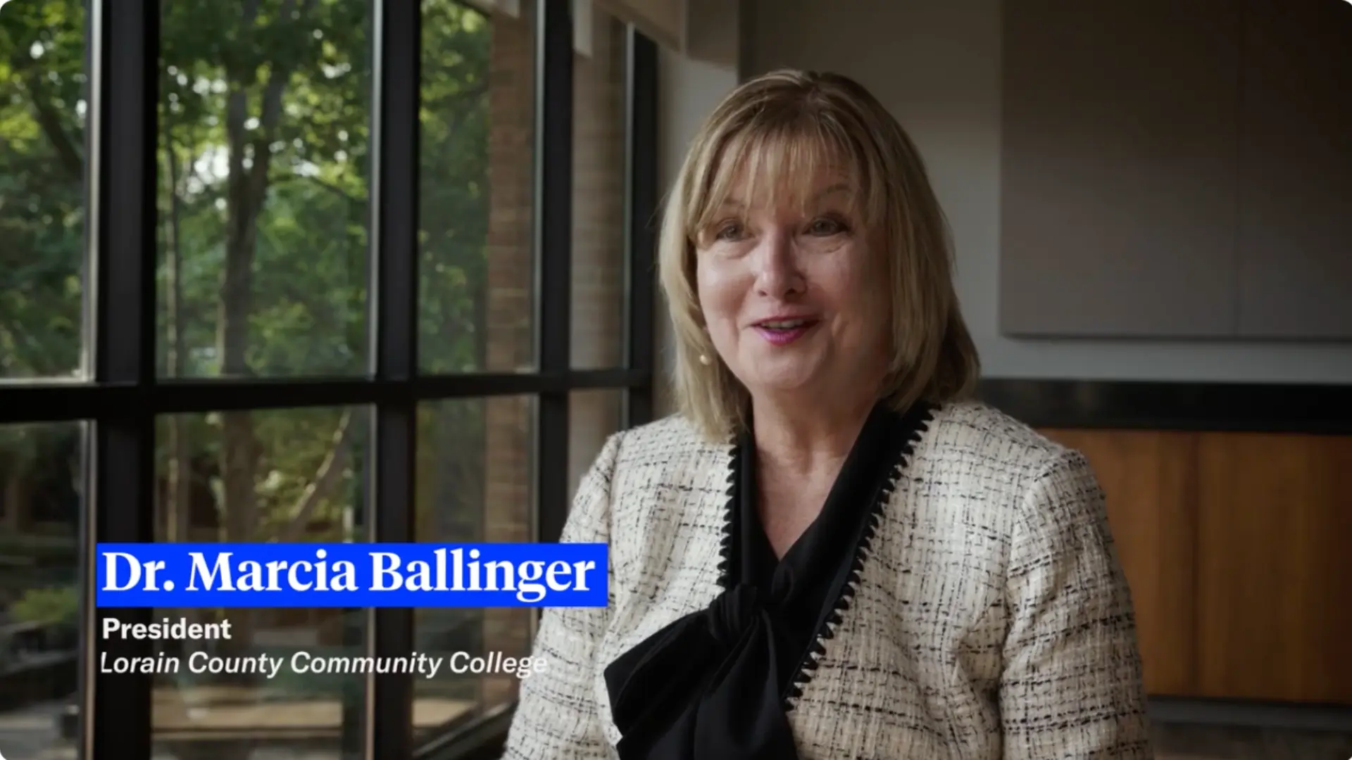 Dr. Marcia Ballinger, President of Lorain County Community College, sits smiling by large windows overlooking greenery.