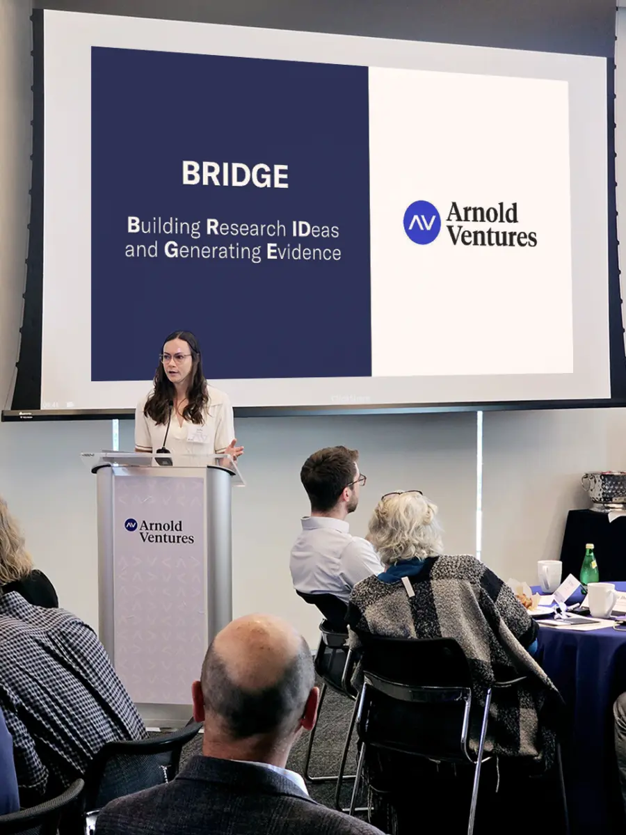 A speaker presents at an AV BRIDGE event, while an engaged audience listens at round tables.