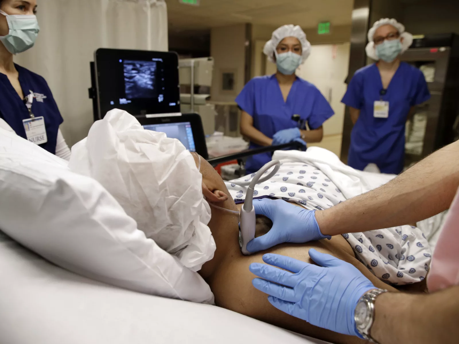 A healthcare team performs an ultrasound on a patient lying in bed, with staff dressed in scrubs and protective gear.