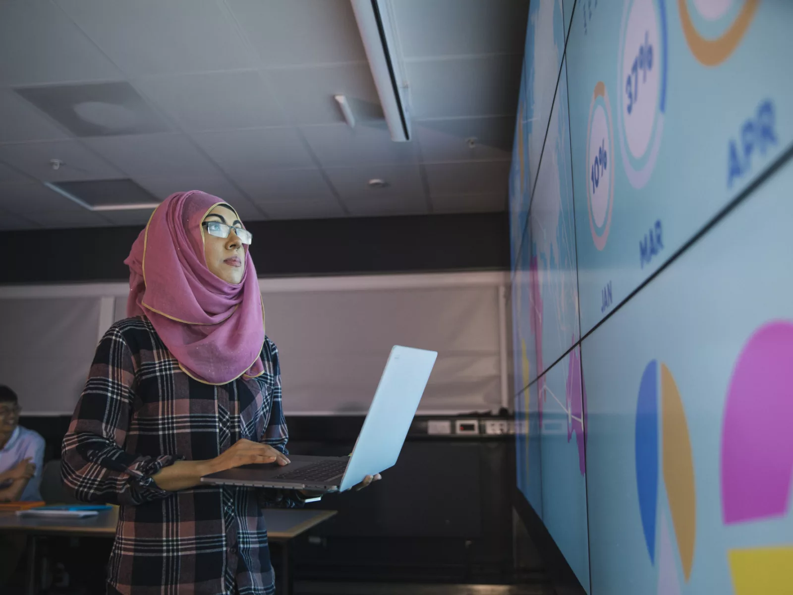 College-age woman holding laptop looks at smartboard that displays graphs in classroom.