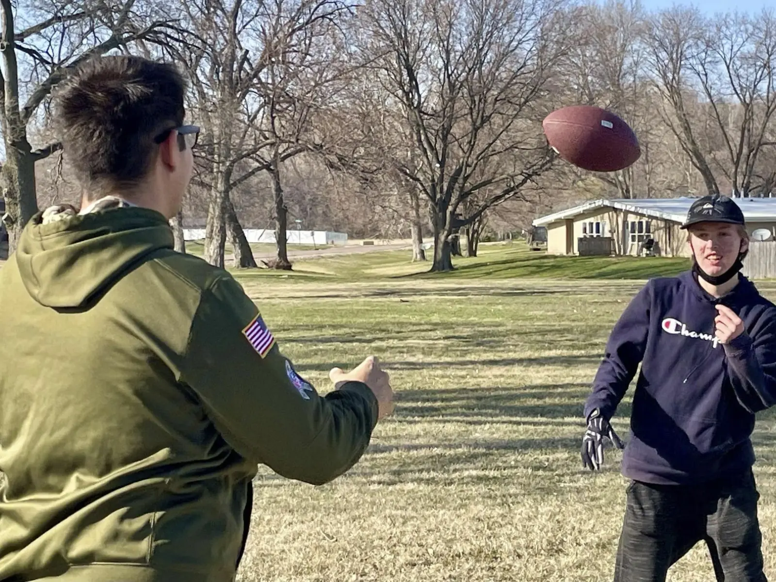 Two young men play catch with a football in a grassy field, surrounded by bare trees and suburban homes in the background.