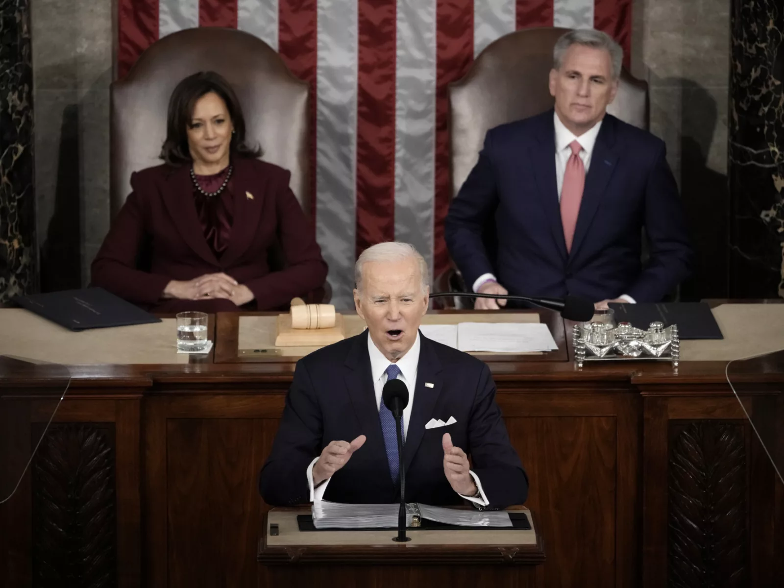 President Joe Biden speaks into a microphone and gestures as Vice President Kamala Harris and Speaker of the House Kevin McCarthy are seated behind him.