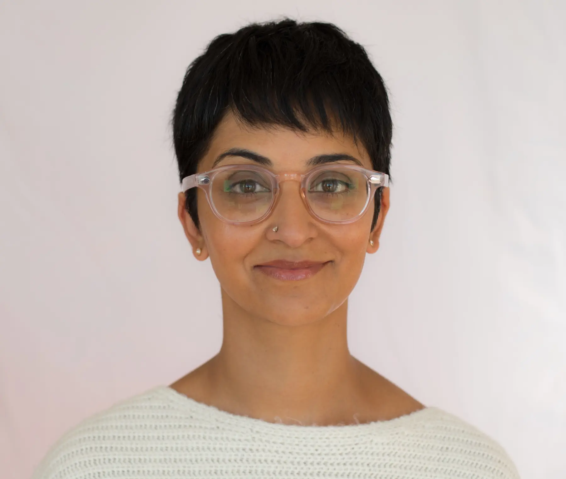 A smiling person with short, dark hair and glasses, wearing a cozy white sweater, stands against a soft, light background.