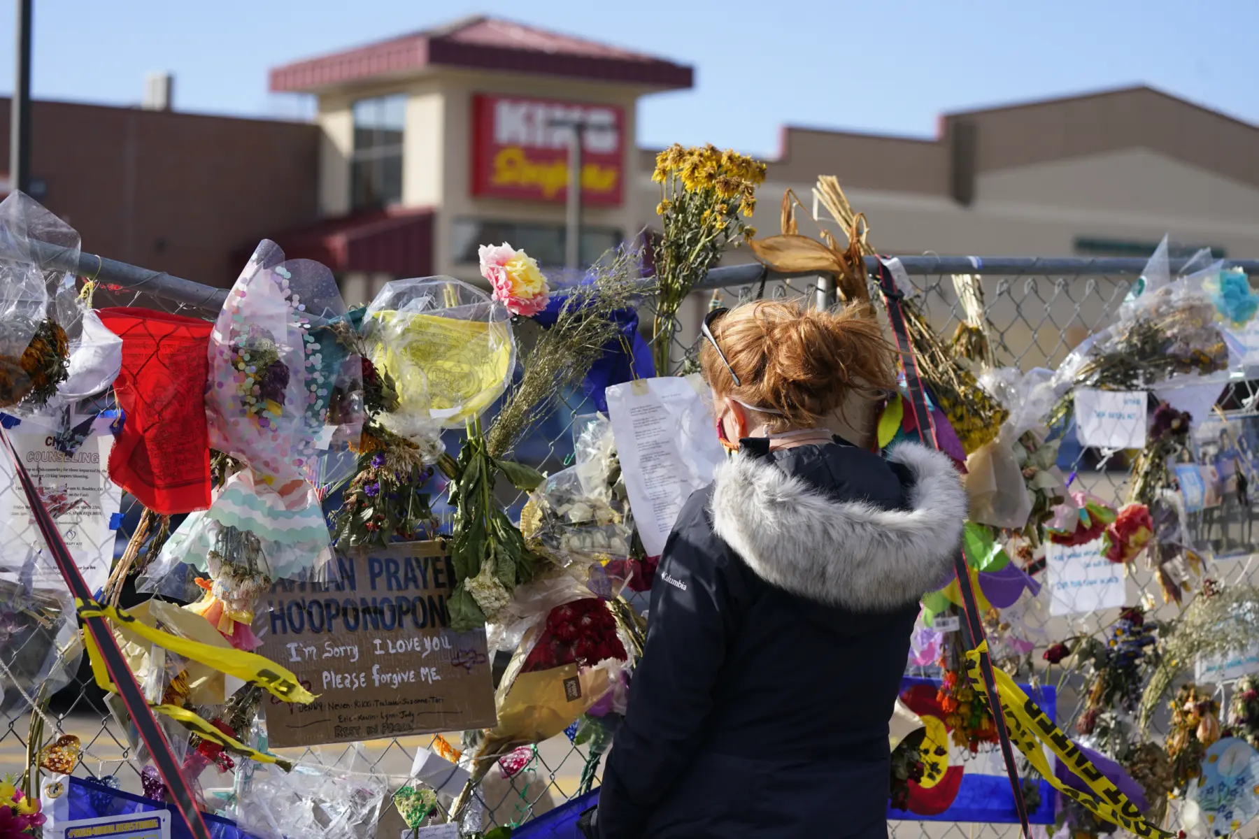 A woman stands before a colorful memorial on a fence, adorned with flowers, notes, and heartfelt messages of love and remembrance.