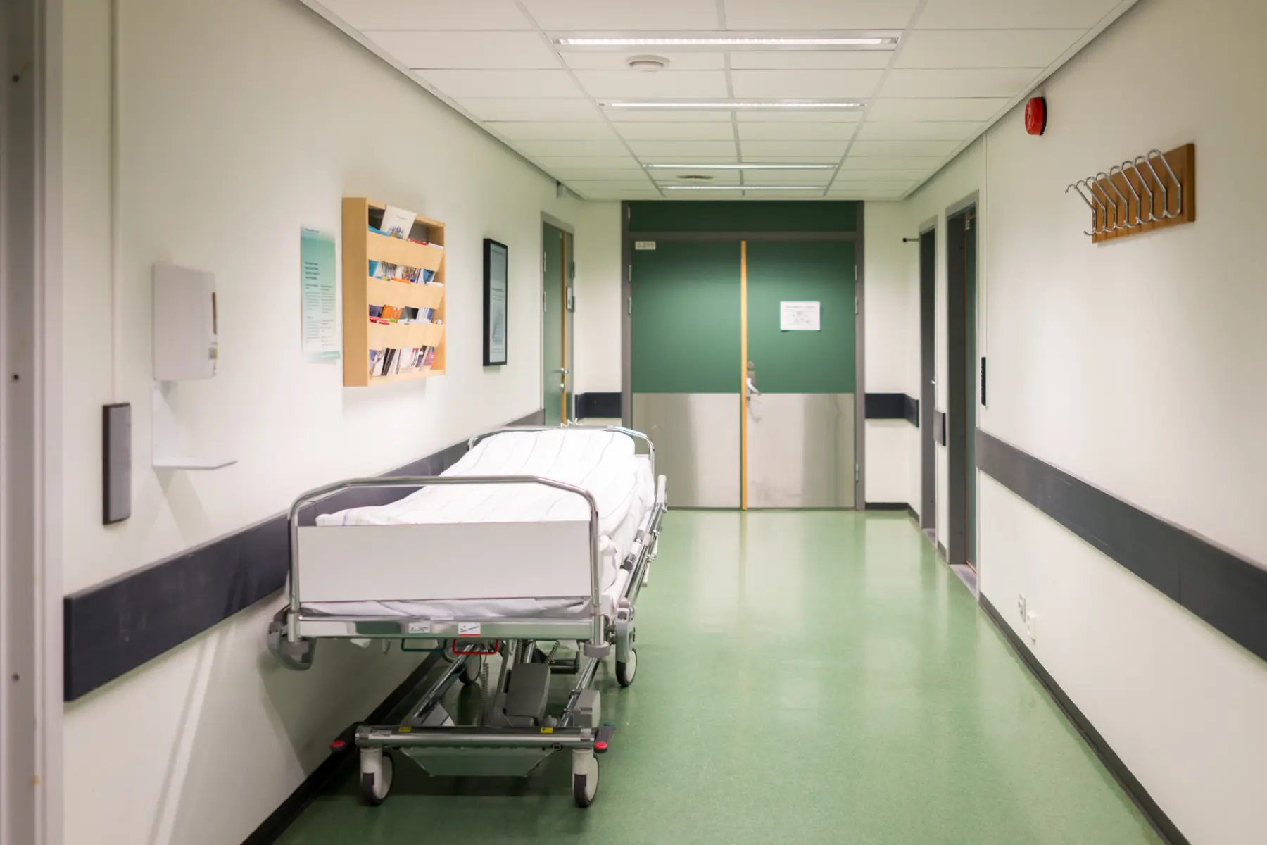 A narrow hospital corridor features a wheeled bed, wooden brochure holders, and green double doors at the far end, with neutral walls.